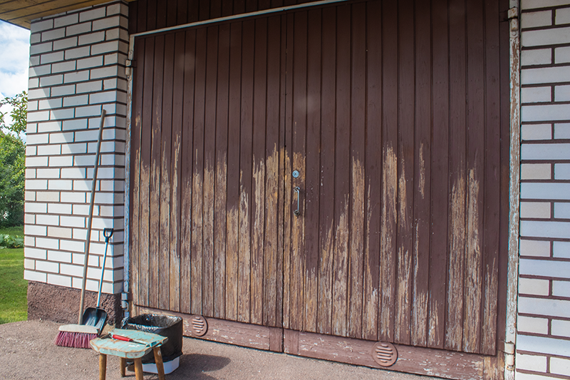 Garagedeuren van hout met gedeeltelijk afgebladderde verf en voorbereide schoonmaakbenodigdheden ernaast.