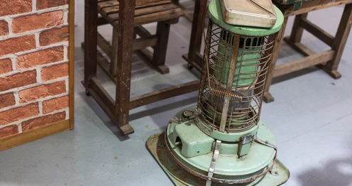 Een vintage petroleumkachel die kachelbrandstof gebruikt, geplaatst op een grijze vloer naast een houten tafel en stoel. De kachel is groen met metalen beschermroosters en staat op een metalen plaat.