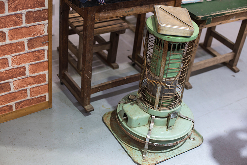 Een vintage petroleumkachel die kachelbrandstof gebruikt, geplaatst op een grijze vloer naast een houten tafel en stoel. De kachel is groen met metalen beschermroosters en staat op een metalen plaat.