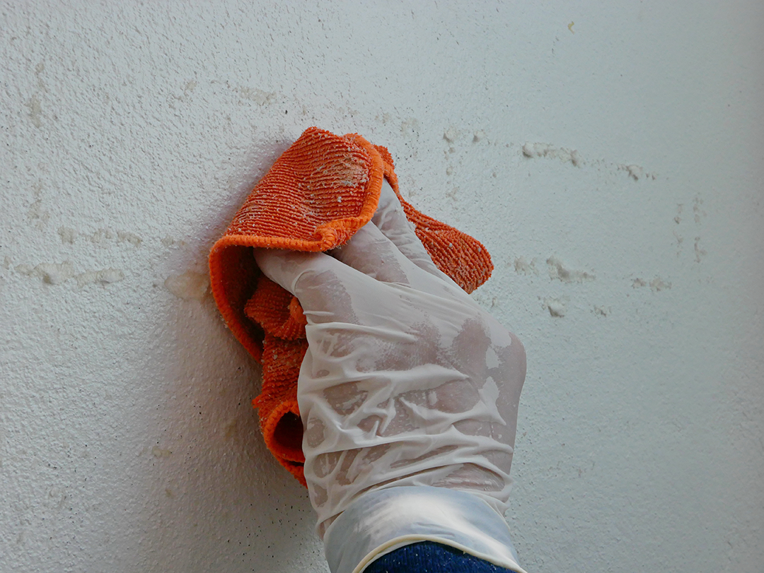 Een hand met een plastic handschoen gebruikt een oranje doek om lijmresten van een wit muuroppervlak te verwijderen.